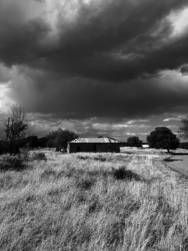 Black and white landscape photograph featuring a small rustic building situated in a grassy field under a dramatic, dark cloudy sky.