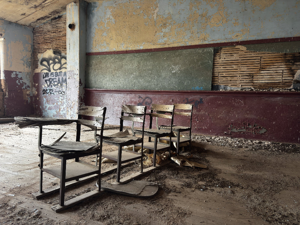 A set of old wooden classroom desks and chairs in a derelict, abandoned room with peeling paint, a chalkboard, graffiti, and a dusty concrete floor.