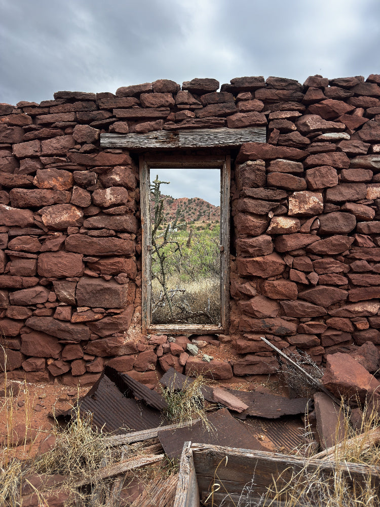 A vertical stone wall made of reddish-brown rocks, featuring a rectangular window looking out onto a landscape, with dry grass and rusted metal debris on the right against a cloudy sky.