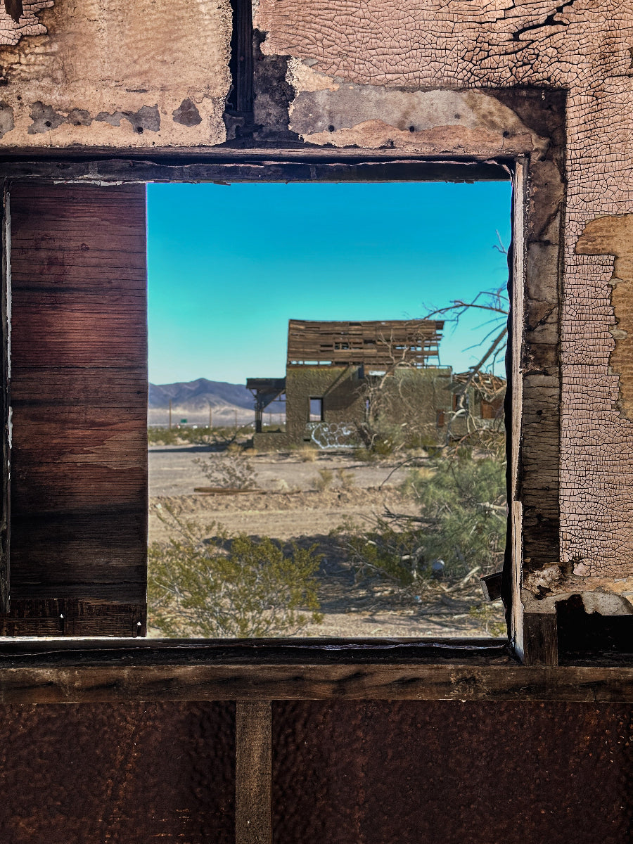 A photograph looking out from a window of an abandoned structure, depicting a desert landscape with a dilapidated building in the background.