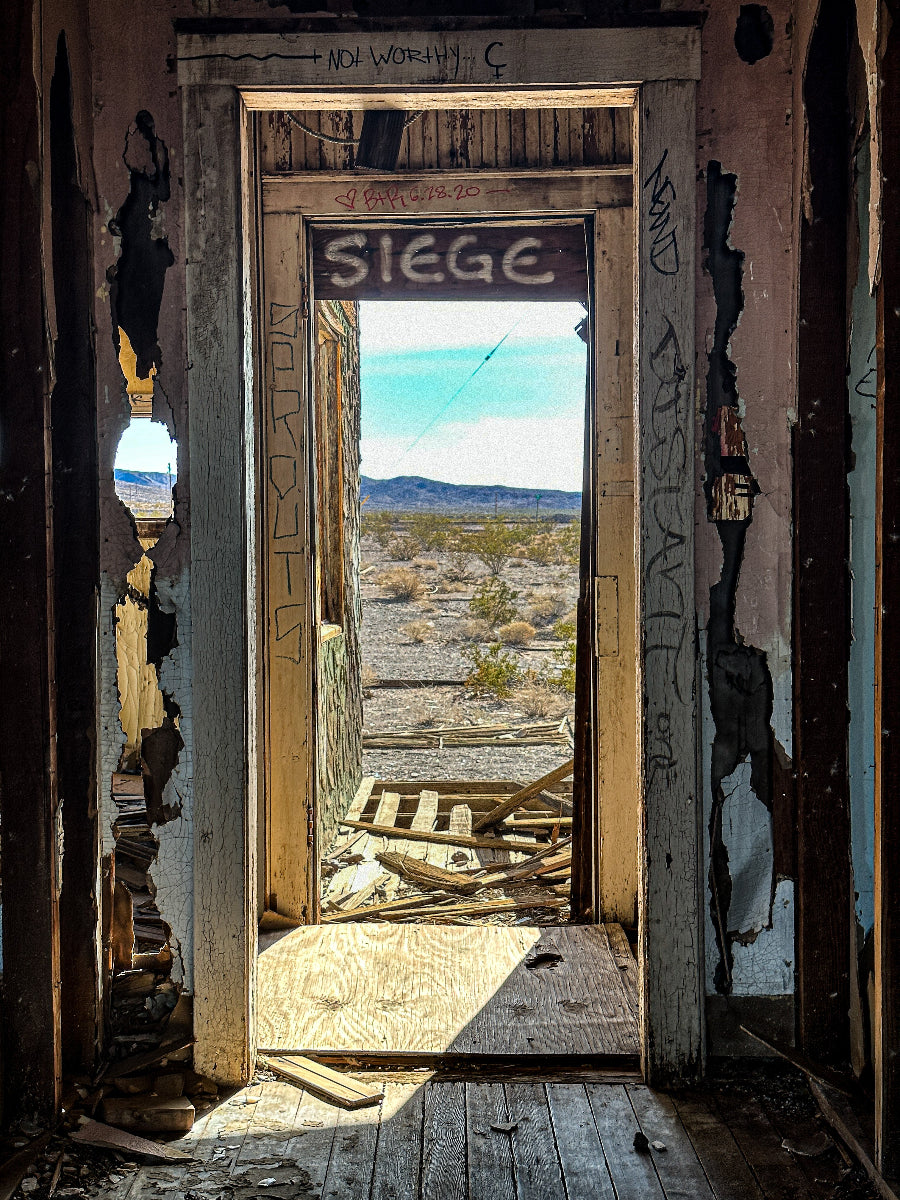 Photograph showing a dilapidated structure with the word 'SIEGE' carved on the lintel, leading to a desert landscape.