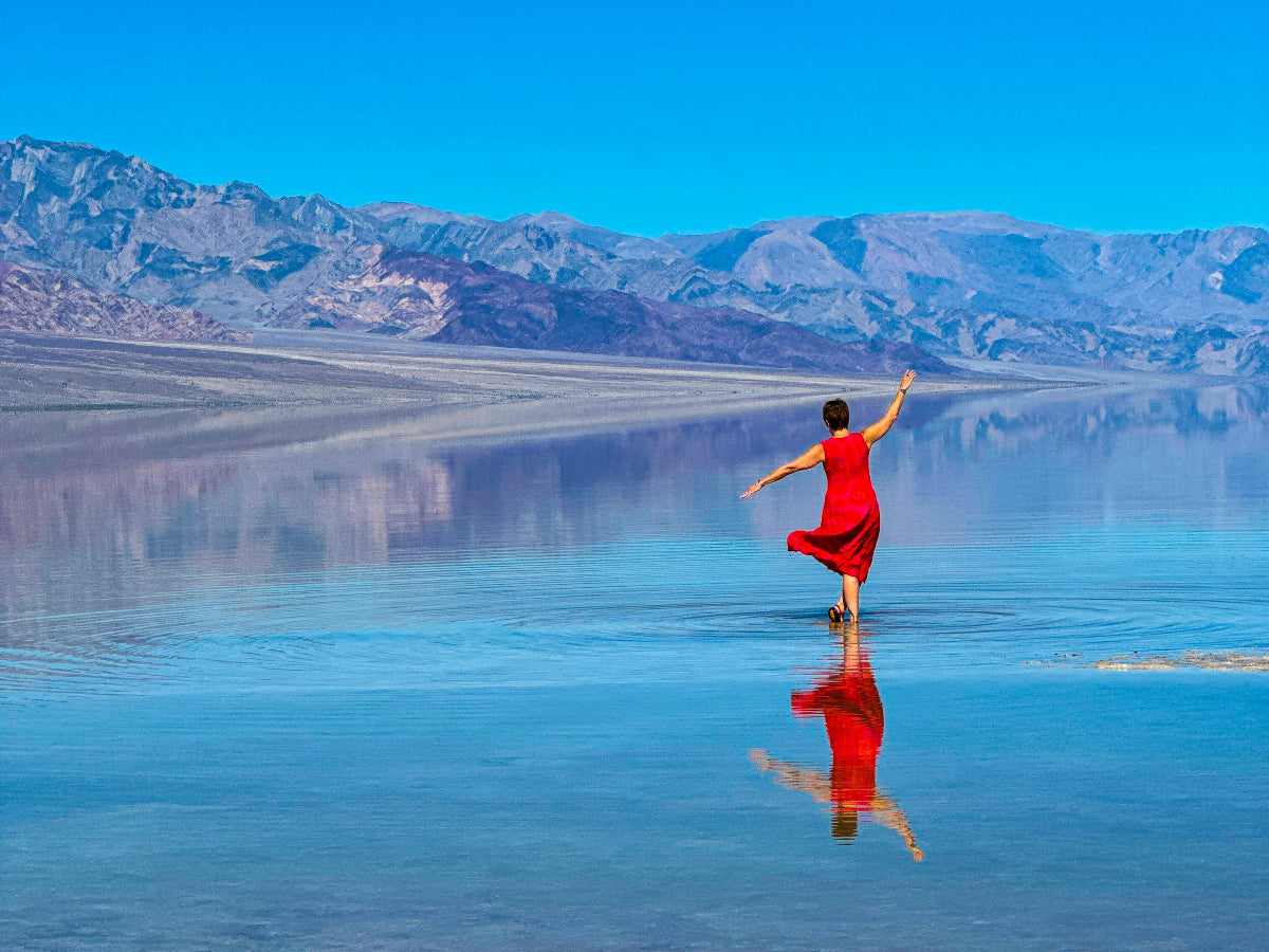 Person in a red dress standing on a reflective water surface with mountains in the background