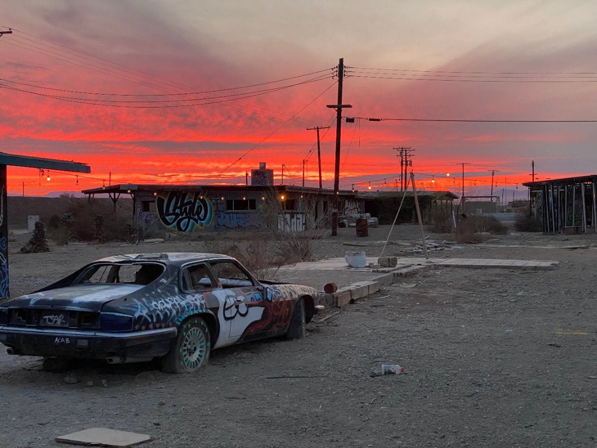 A photograph taken at Bombay Beach, Salton Sea, California, featuring an abandoned car with graffiti and a sunset in the background.