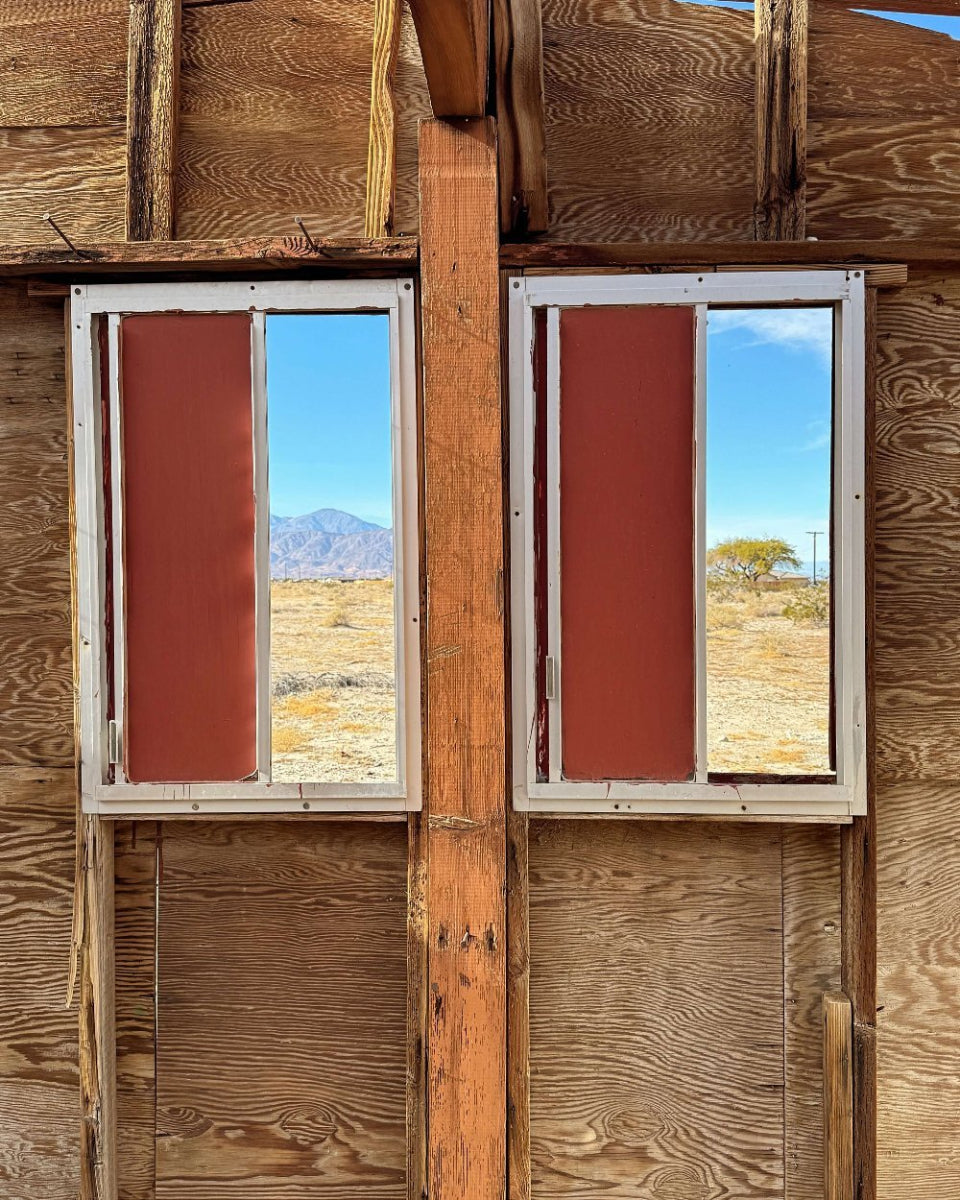 Two windows with red shutters frame a desert landscape in the background, set against a wooden structure that appears burnt on the sides.