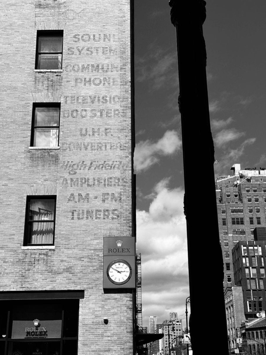 Black and white photo of a Rolex clock on the side of a building with vintage electronics store lettering.