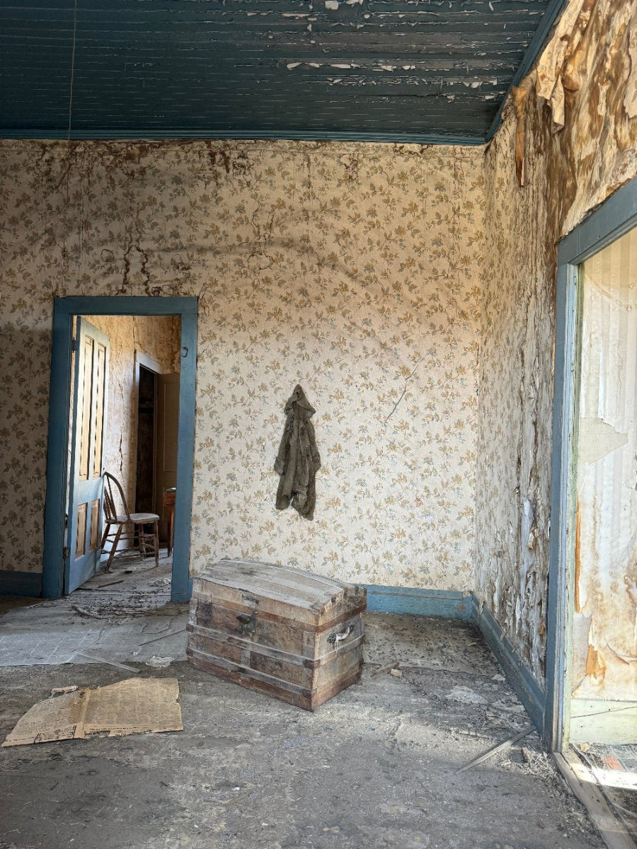 Interior of an abandoned room in a state of decay, with walls covered in patterned wallpaper, a wooden trunk, and a hanging coat rack.