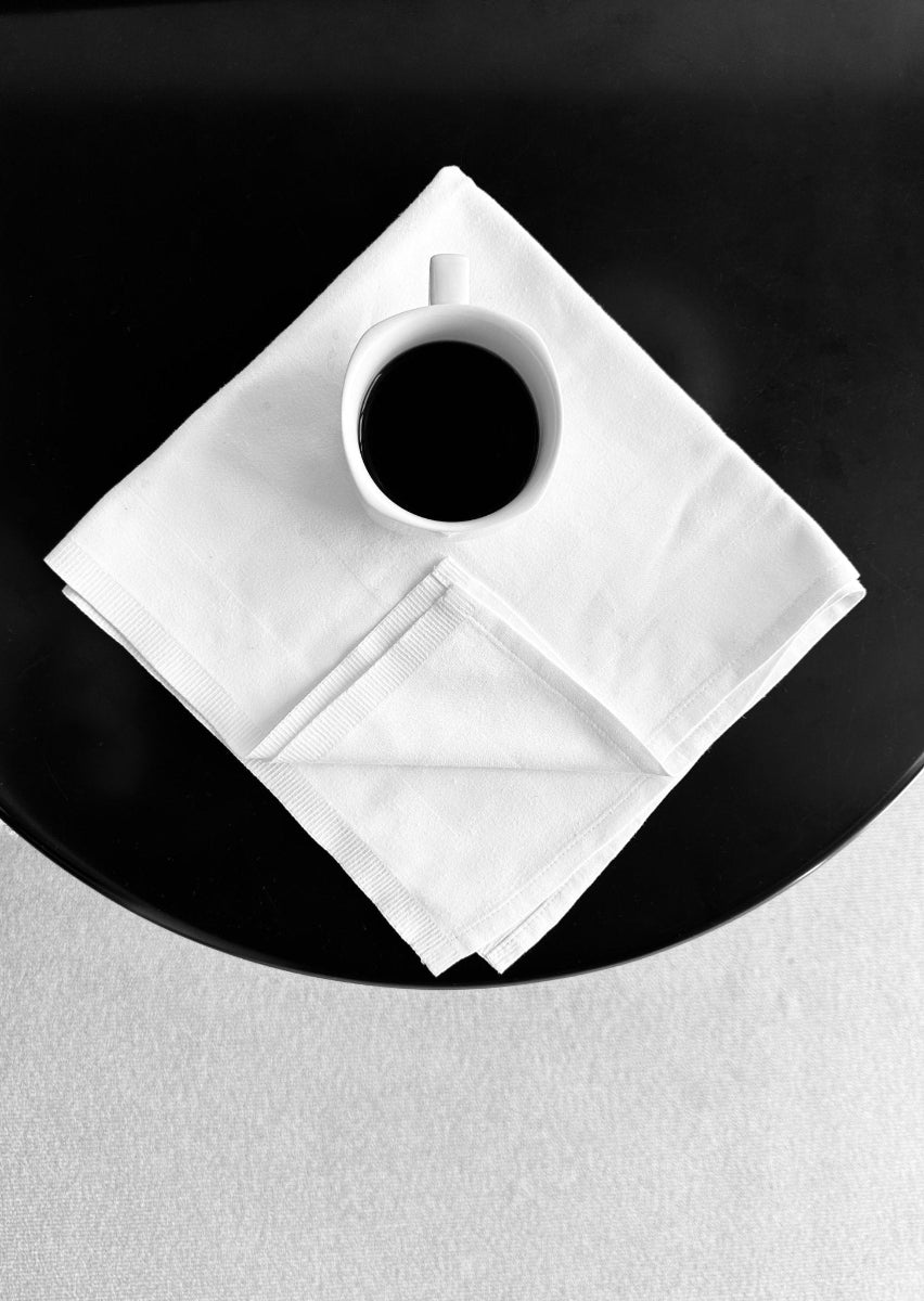 A black and white photograph of a white coffee cup filled with coffee, placed on a neatly folded white cloth napkin, on a black circular table.