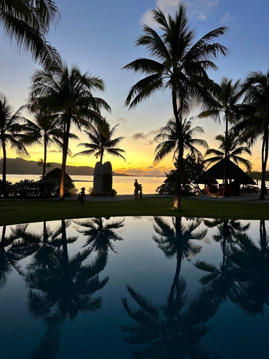 A photo of a Tahiti sunset reflecting in a pool, with silhouettes of palm trees and a person in the distance.