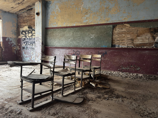 A set of old wooden classroom desks and chairs in a derelict, abandoned room with peeling paint, a chalkboard, graffiti, and a dusty concrete floor.