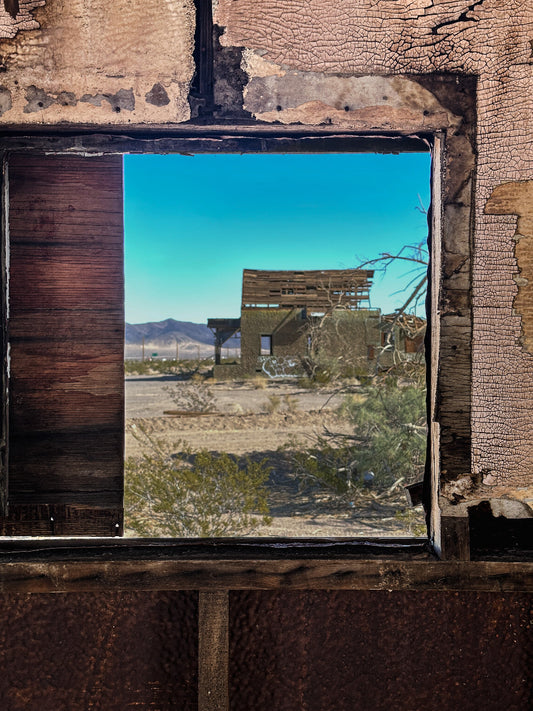 A photograph looking out from a window of an abandoned structure, depicting a desert landscape with a dilapidated building in the background.