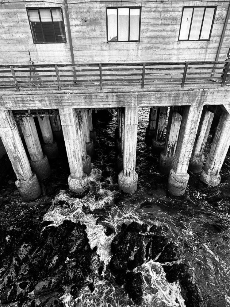 Black and white photograph of a weathered wooden pier structure with large support columns over churning ocean water and dark rocks.