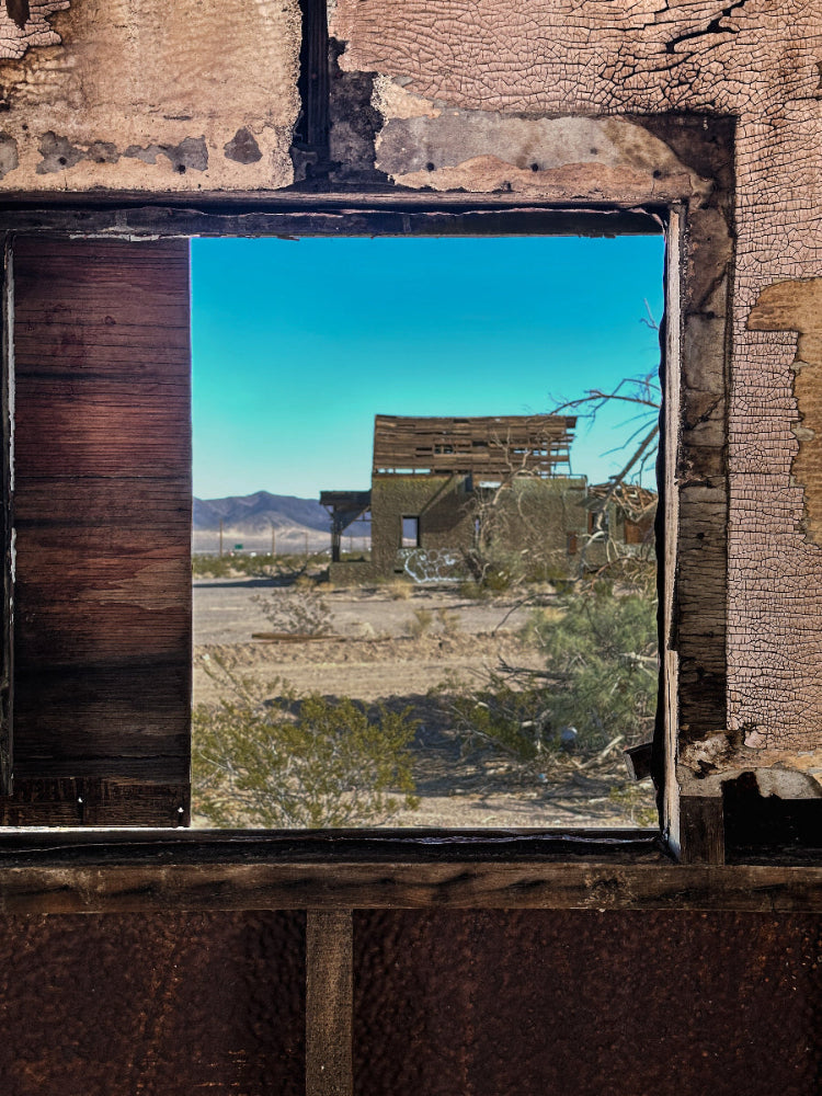 A photograph looking out from a window of an abandoned structure, depicting a desert landscape with a dilapidated building in the background.