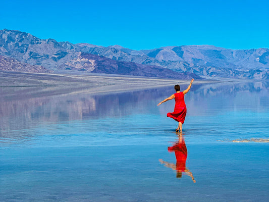 Person in a red dress standing on a reflective water surface with mountains in the background